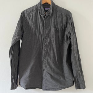 Peut inclure: Une chemise à manches longues boutonnée de couleur gris foncé avec une poche poitrine et une étiquette Patagonia sur le côté gauche.