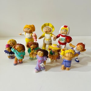 May include: A collection of nine Cabbage Patch Kids figurines. The figurines are all different colors and have different outfits. Some of the figurines are holding accessories, such as a doll, a football, and an ice cream cone.