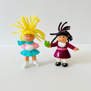 Peut inclure: Deux poupées Cabbage Patch Kids habillées en vêtements d'hiver. Une poupée a des cheveux en laine jaune et porte un pull rose, une jupe bleue et des patins à glace blancs. L'autre poupée a des cheveux en laine brune et porte une robe bordeaux et des chaussures noires. La poupée aux cheveux jaunes tient une boule de neige blanche. La poupée aux cheveux bruns tient un cadeau vert.