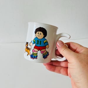 Puede incluir: Taza de cerámica blanca con una ilustración de dibujos animados de un niño con patines y un perro. La camiseta del niño tiene el número 31. La taza tiene un asa curva y un acabado brillante.