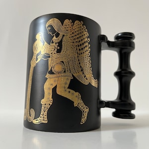May include: Black ceramic mug with a gold-colored illustration of a figure pouring water from a pitcher. The figure is wearing a robe and has wings. The mug has a black handle.