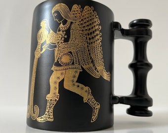 Pode incluir: Caneca de cerâmica preta com uma ilustração dourada de uma figura despejando água de um jarro. A figura está vestindo uma túnica e tem asas. A caneca tem uma alça preta.