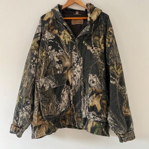 May include: A green, brown, and white camouflage hooded jacket with a zipper closure and a Buck Wear logo on the front. The jacket has a large pocket on the left side and a drawstring at the bottom.