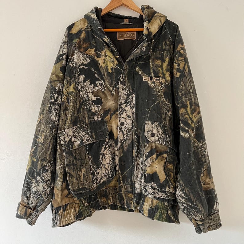May include: A green, brown, and white camouflage hooded jacket with a zipper closure and a Buck Wear logo on the front. The jacket has a large pocket on the left side and a drawstring at the bottom.