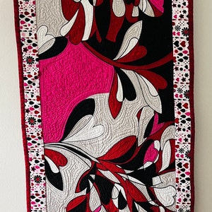 Peut inclure: Une tenture murale matelassée avec un motif abstrait moderne composé de feuilles stylisées noires, blanches, rouges et roses. La bordure est en tissu rose et blanc avec des cœurs et des fleurs.