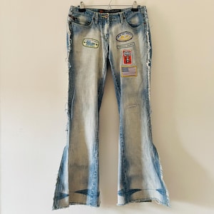 Peut inclure: Jean en denim bleu clair avec une coupe évasée. Le jean comporte plusieurs écussons, dont un drapeau américain et du texte. Les bords effilochés ajoutent un aspect usé. Le jean est suspendu à un cintre en métal.
