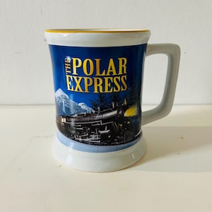 Op de afbeelding: Een keramische mok met een wit handvat en voet, met een blauwe achtergrond met de woorden "THE POLAR EXPRESS" in gouden letters. De mok toont een trein en besneeuwde bergen.