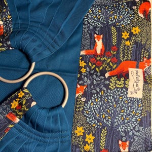 Pode incluir: Um sling de argolas azul-petróleo com argolas prateadas e um painel de tecido com tema de floresta. O tecido apresenta raposas laranja, flores amarelas e folhagem verde sobre um fundo azul marinho. A marca "sewfunkyea" é visível.