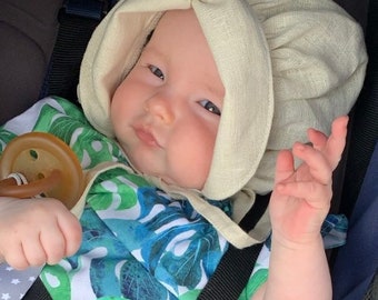 Könnte beinhalten: Ein Baby trägt eine weiße Leinenhaube und einen grünen und blauen Tropen-Druck-Strampler. Das Baby sitzt in einem Autositz und hat einen Schnuller im Mund.