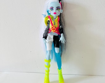 May include: A doll with blue skin, black and white hair, and a neon yellow and pink outfit. The doll has a black mesh top, a yellow vest, and white and black shorts. The doll is wearing neon yellow boots.
