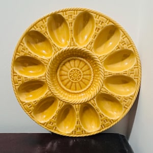 Puede incluir: Una fuente redonda de cerámica amarilla para servir ostras con 12 hendiduras ovaladas y un pozo central circular. La fuente presenta un diseño de cesta tejida alrededor de los bordes y el centro, creando una apariencia texturizada. El color es un amarillo dorado cálido.