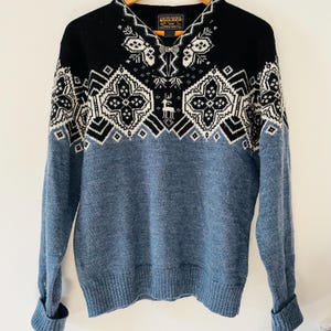 May include: A Woolrich V-neck sweater with a black upper section and blue lower section. The sweater features a geometric pattern in white and black, with a deer motif. The sleeves are cuffed, and the sweater has a tag at the neckline.