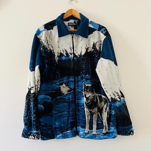 May include: A long-sleeve zip-up fleece jacket featuring a blue and white winter landscape print with a wolf standing in the foreground. The jacket has a solid blue collar, a full-length black front zip, and a standard fit with long sleeves.