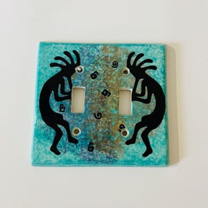 May include: A square light switch cover with a turquoise and brown mottled background. Two black Kokopelli figures flank the two switch openings. The cover has a glossy finish.