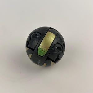 Op de afbeelding: Een zwart en goudkleurig bolvormig speelgoed met een groene accentkleur. Het speelgoed heeft een segmentontwerp met zichtbare scharnieren en details. Het gouden gedeelte heeft een groen grasachtig ontwerp. Het speelgoed ligt op een witte ondergrond.