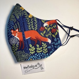 May include: A blue fabric face mask with a fox and floral pattern. The mask has adjustable ear loops with black toggles. The mask has a white tag with the text "sewfunky.ca" and a dragonfly.