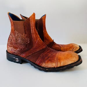 May include: A pair of brown leather ankle boots with a decorative floral design on the side. The boots have a distressed look and a chunky heel.