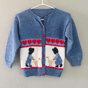 May include: A blue knitted cardigan with a round neck and long sleeves. The sweater features a white panel with two figures and geese, red hearts, and red buttons. The cardigan is hanging on a white hanger.