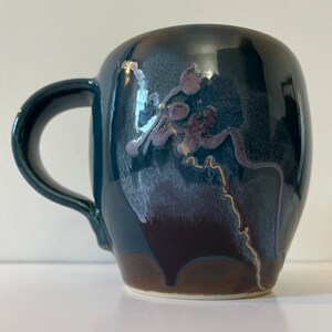 Peut inclure: Une tasse en céramique bleu foncé avec un design unique présentant un motif abstrait blanc et violet avec des accents dorés. La tasse a un corps arrondi et une anse.