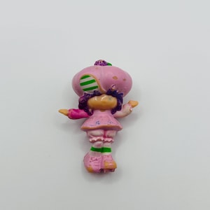 Peut inclure: Petite figurine colorée représentant un personnage avec un chapeau rose en forme de champignon à rayures vertes. Le personnage porte une robe rose à petits pois bleus, des cheveux violets et des chaussettes rayées vertes et roses. Sur fond blanc.