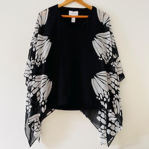 May include: Black V-neck top with white butterfly wing designs on the sleeves. The top is hanging on a wooden hanger against a white wall. The top has a sheer, flowing design.