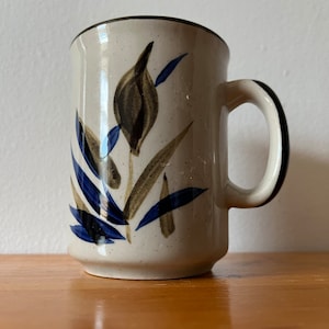 May include: A white ceramic mug with a brown rim and handle. The mug features a floral design with brown and blue leaves and a brown flower.