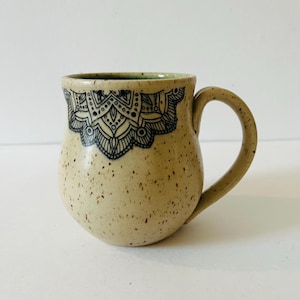 Puede incluir: Una taza de cerámica beige moteada con un diseño de mandala en blanco y negro alrededor del borde.