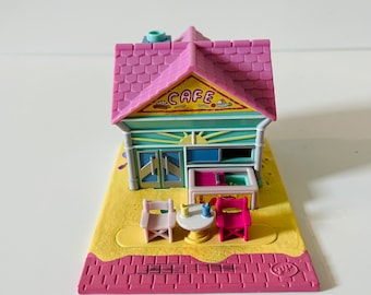 May include: A pink and blue plastic toy cafe playset with a yellow awning that says "CAFE" and a table and chairs outside.