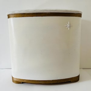 Könnte beinhalten: Ein weißer Vintage-Schrank mit einer strukturierten, gerippten Oberfläche. Der Schrank hat goldfarbene Zierleisten an Ober- und Unterseite sowie ein silbernes Sternemblem auf der Vorderseite. Die Oberseite scheint aus hellem Stein oder Marmor zu sein.