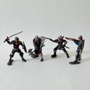 Könnte beinhalten: Vier Miniaturritterfiguren in Kampfposen. Die Ritter sind in detaillierter Rüstung mit Schwertern, Äxten und Armbrüsten gekleidet. Das Farbschema umfasst Schwarz, Rot, Silber und Gold und erzeugt ein dynamisches und historisches Thema.