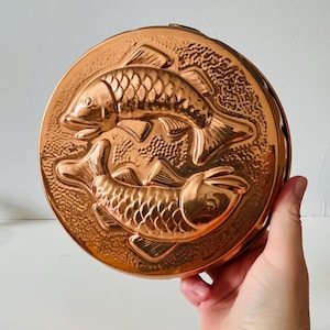 Puede incluir: Un molde redondo de color cobre con un relieve detallado de dos peces. Los peces están orientados en direcciones opuestas, con escamas y aletas claramente definidas. El molde tiene un acabado brillante y es probablemente para uso decorativo o culinario.