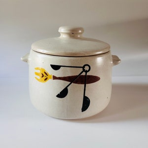 May include: A white ceramic crock with a lid, featuring a brown, black, and yellow design of a spoon, fork, and spatula.