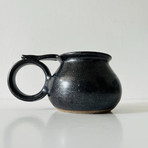 Peut inclure: Une tasse en céramique noire tachetée avec un corps arrondi et bombé, dotée d'une grande anse circulaire sur le côté. La base de la tasse présente un bord en argile brune non émaillée, contrastant avec la finition sombre et brillante.