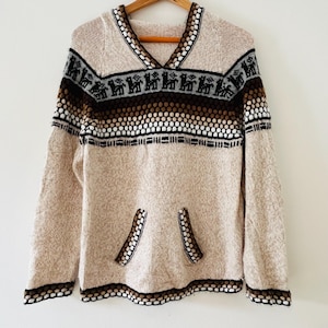 May include: A beige knit hoodie with a V-neck and a kangaroo pocket. The sweater features a patterned design in black, brown, and white around the neckline and chest. The cuffs and hem are also trimmed with a patterned border.