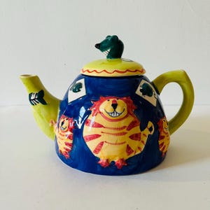 Könnte beinhalten: Eine blaue Teekanne aus Keramik mit einem grünen Mausdeckel. Die Teekanne ist mit gelben, roten und orangefarbenen gestreiften Katzen verziert.