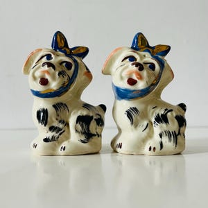 May include: A pair of ceramic dog figurines. The dogs are white with black spots, blue bows, and blue and gold accents. The figurines have a vintage aesthetic and are likely salt and pepper shakers.