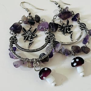 May include: A pair of handcrafted earrings featuring silver hoops adorned with amethyst chips and fairy charms. Each earring has a dangling mushroom charm with a burgundy cap and white stem. The earrings are designed for pierced ears.