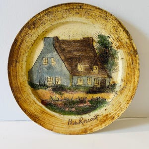 May include: A round ceramic plate with a textured, beige-toned rim. The center features a relief illustration of a house with a blue facade and brown roof, surrounded by green trees and a field. The text H du Roscouet is inscribed at the bottom.