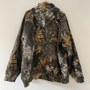 May include: A hooded jacket with a camouflage pattern in shades of brown, green, and white. The jacket has a drawstring hood and a zipper closure.