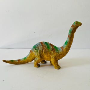 Puede incluir: Un juguete de dinosaurio Braquiosaurio amarillo y verde con cuello y cola largos. El juguete tiene marcas marrones y verdes en la espalda y una cabeza verde. El dinosaurio est&aacute; de pie sobre sus cuatro patas.