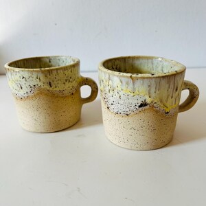 May include: A pair of handmade ceramic mugs with a speckled, textured finish. The mugs feature a beige base, with a yellow and brown glaze towards the top. Each mug has a small, rounded handle, ideal for tea or coffee.