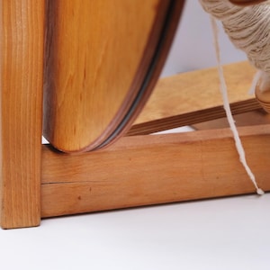 May include: A wooden spinning wheel with a large, curved wooden wheel and a spool of white yarn. The wheel is made of light-coloured wood with a dark rim. The yarn is wound around a wooden bobbin.