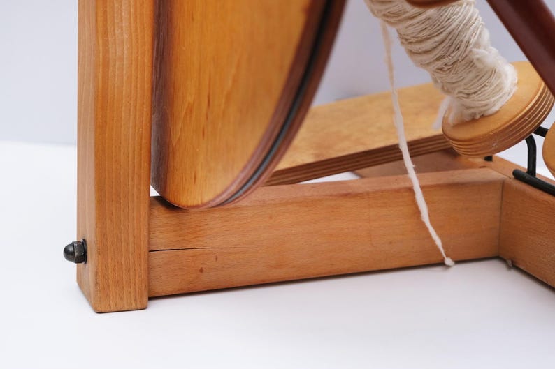 May include: A wooden spinning wheel with a large, curved wooden wheel and a spool of white yarn. The wheel is made of light-coloured wood with a dark rim. The yarn is wound around a wooden bobbin.