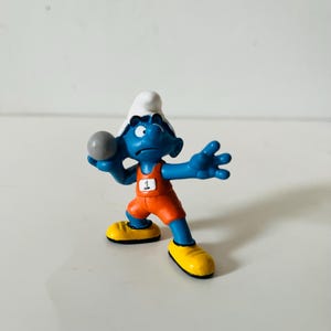 May include: A blue Smurf figurine in an orange athletic uniform, holding a gray ball. The figurine has a white hat and yellow shoes. The number 1 is visible on the chest of the uniform. The Smurf is in a throwing pose.
