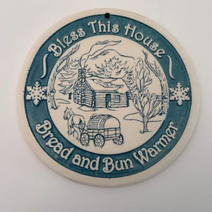 May include: Round ceramic bread and bun warmer with a blue border and a cream-colored center. The center features a cabin scene with a horse-drawn wagon and the text "Bless This House Bread and Bun Warmer."