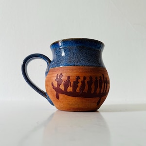 Peut inclure: Une tasse en céramique faite à la main avec un bord et une anse émaillés bleus. Le corps de la tasse est de couleur beige avec une illustration rouge foncé de personnages dans un bateau. La tasse est conçue pour les boissons chaudes.