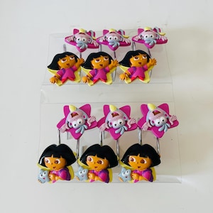 May include: A set of shower curtain hooks featuring a cartoon character with a black bob, orange skin, and a pink outfit. The hooks also include a pink star with a cartoon monkey. The hooks are displayed on a clear plastic stand.