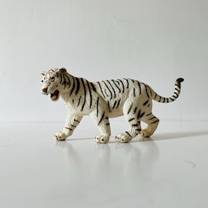 May include: A white tiger figurine with black stripes, depicted in a walking stance with its mouth open. The toy animal features detailed moulded fur texture, four legs, and a long tail, rendered in a realistic style with a cream and black colour palette.