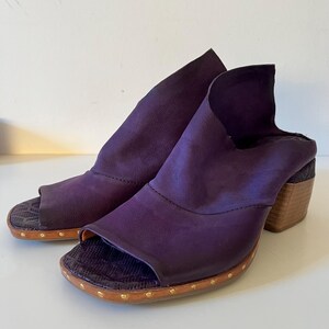 May include: A pair of open-toe, purple leather mules with a wooden heel. The shoes have a textured sole and are adorned with small, gold-colored studs around the base. The design features a unique, folded leather upper.