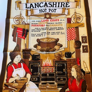 May include: A vintage tea towel with a brown and tan background featuring a recipe for Lancashire Hot Pot. The recipe includes lamb chops, onions, potatoes, and carrots. The image also shows a woman in a red shirt and white apron preparing the dish in a kitchen with a black stove and a fire.